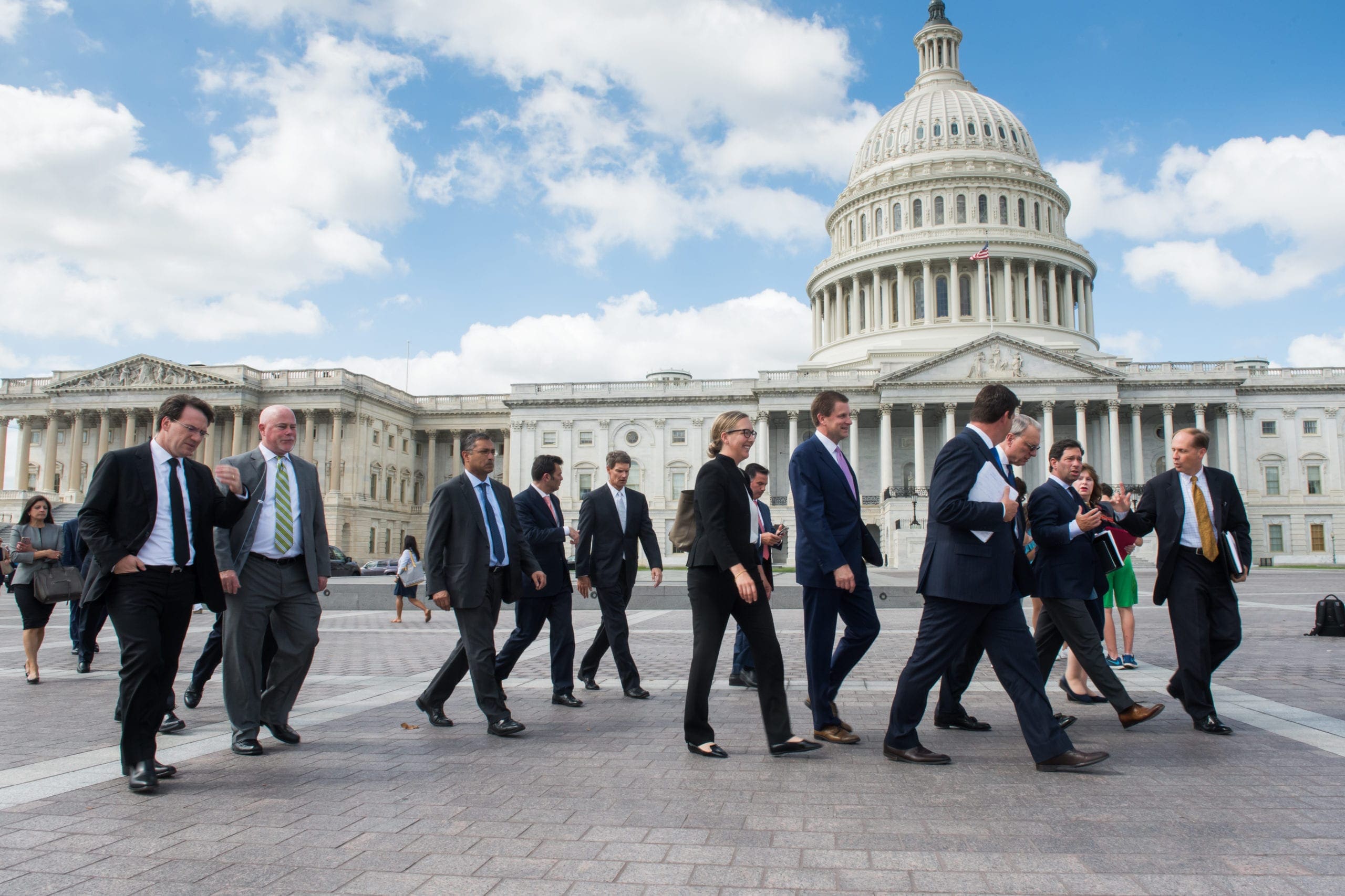A group of men and women in suits walking past the United States Capitol building.